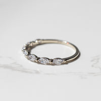A dainty white gold diamond wedding band sitting on a white marble backdrop. The ring as a row of marquise diamonds with a shared single prong between them.