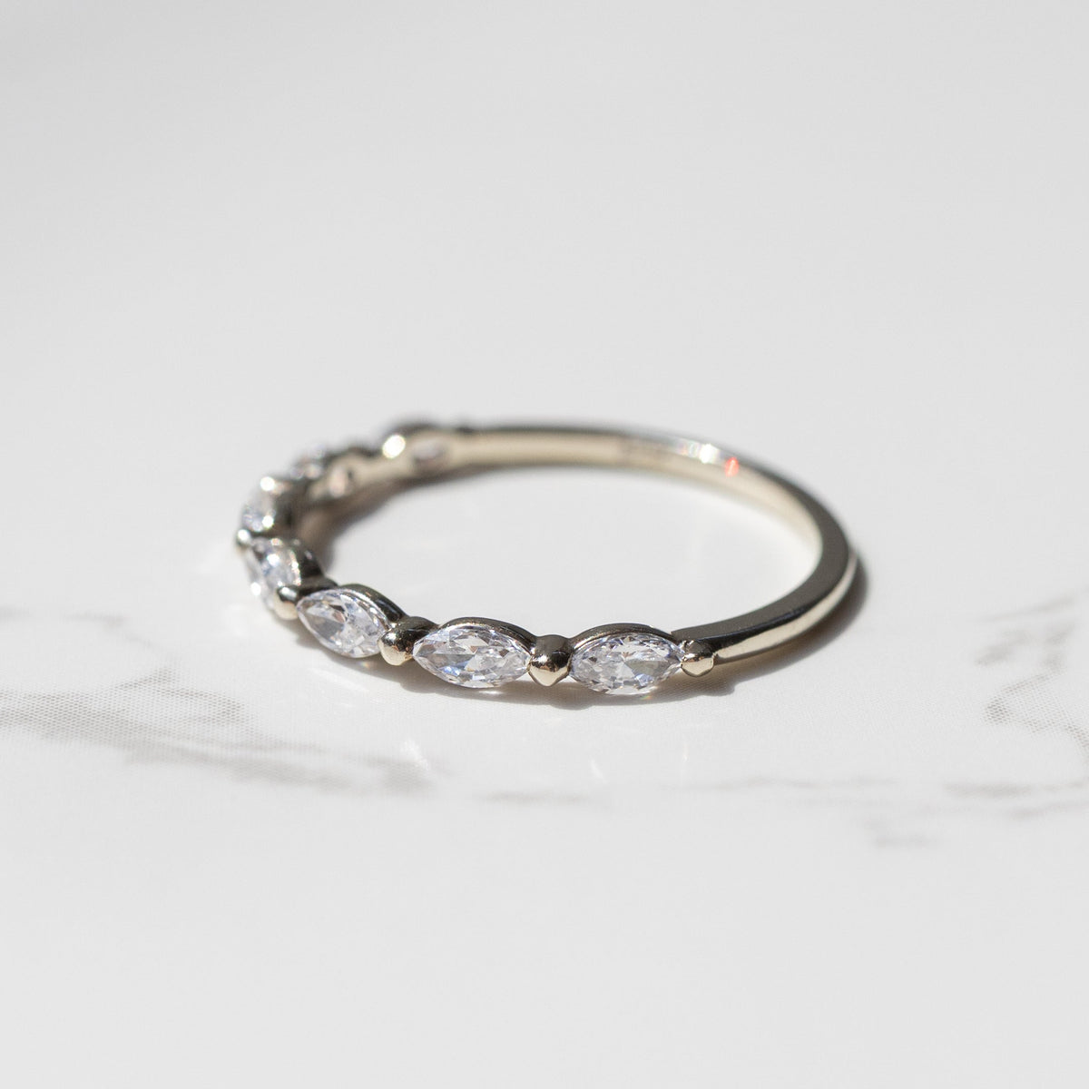 A dainty white gold diamond wedding band sitting on a white marble backdrop. The ring as a row of marquise diamonds with a shared single prong between them.