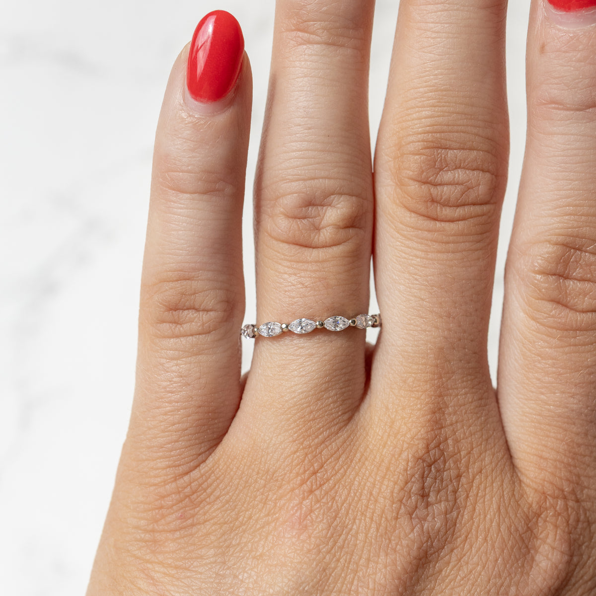 A close-up of a hand wearing a delicate silver band ring with a row of small marquise-cut diamonds, set against a light marble background.