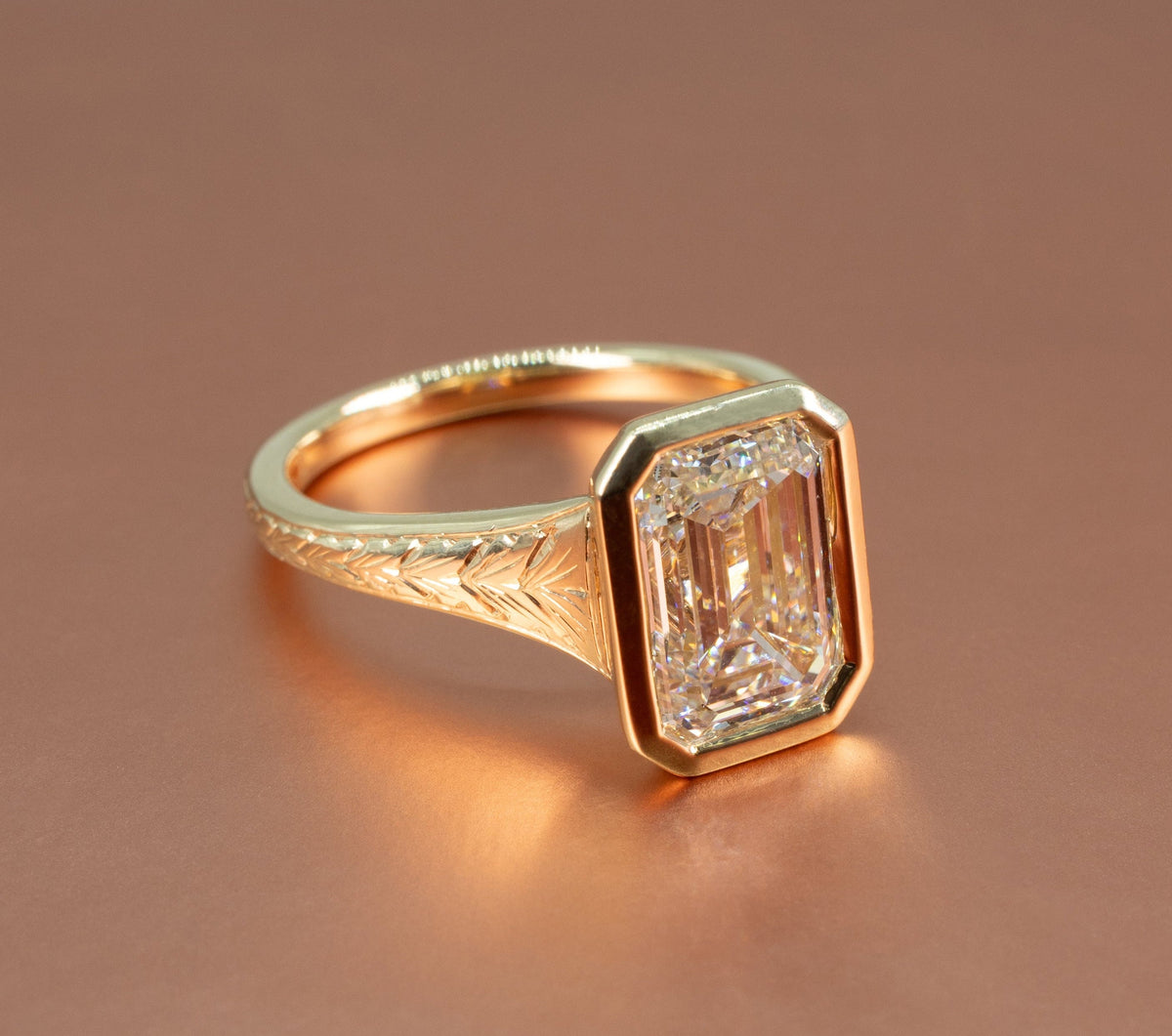 A large yellow gold bezel engagement ring sitting on a rose gold backdrop. The ring has a large emerald cut center diamond and intricate hand engraving on the band.