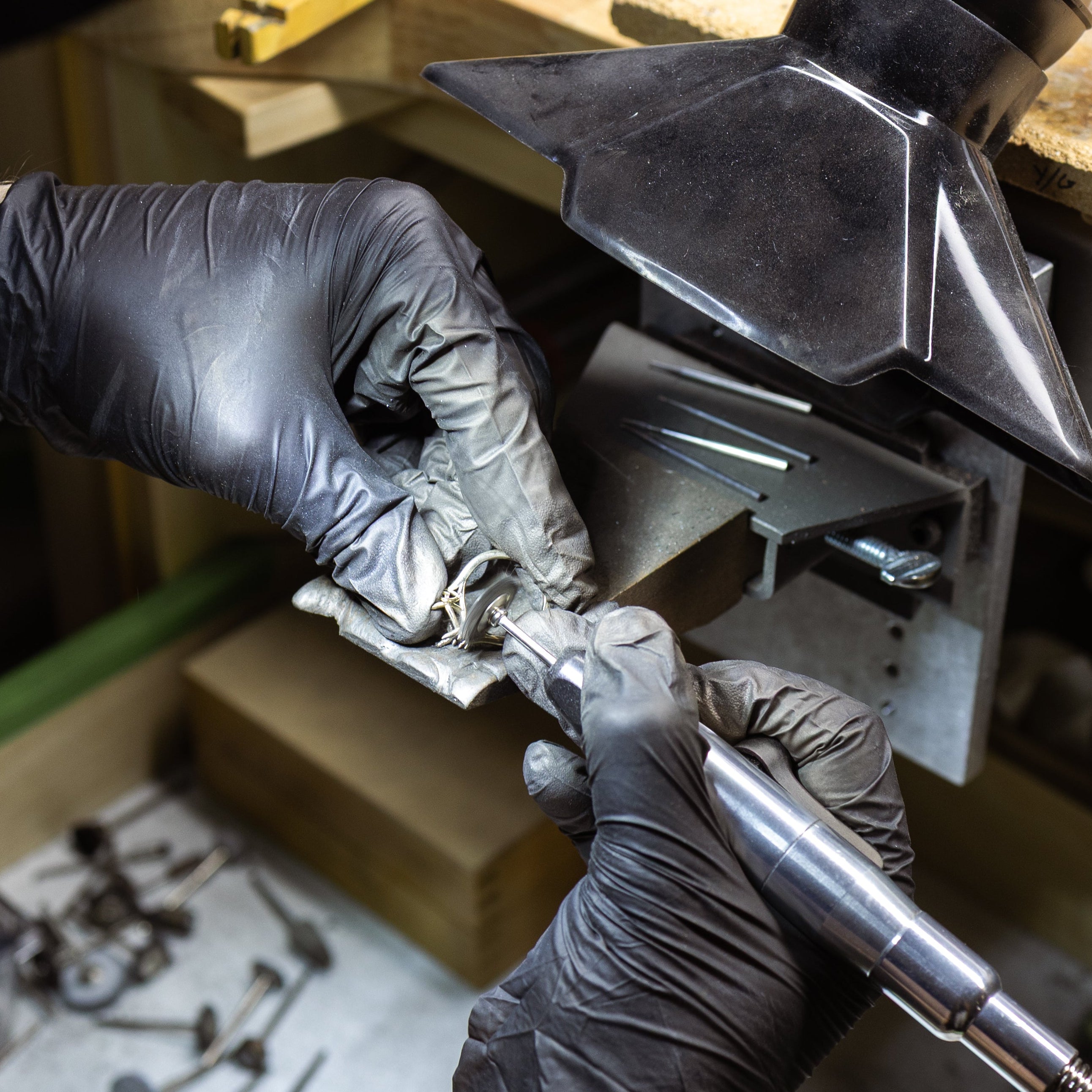 Close-up of a jeweler's hands wearing black nitrile gloves using a metal tube tool under a magnifying lamp on a wooden workbench.