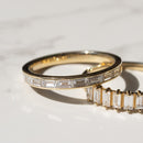 Two gold rings with clear baguette-cut diamonds, one shown in profile and the other partially visible underneath, resting on a white marble surface.
