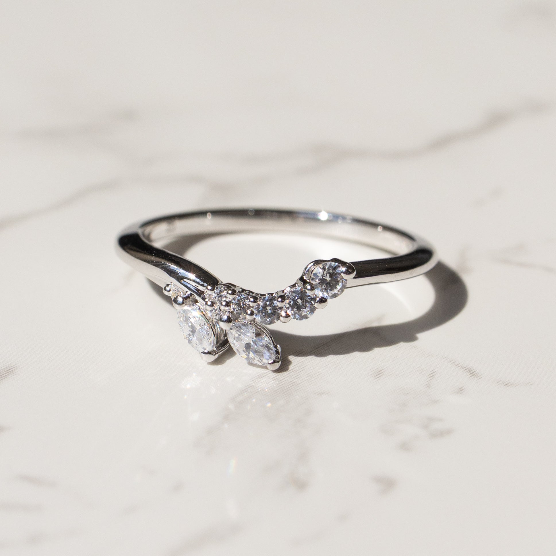 A silver ring featuring a curved, leaf-like cluster of clear marquise and round gemstones, resting on a light marble surface.
