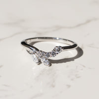 A silver ring featuring a curved, leaf-like cluster of clear marquise and round gemstones, resting on a light marble surface.