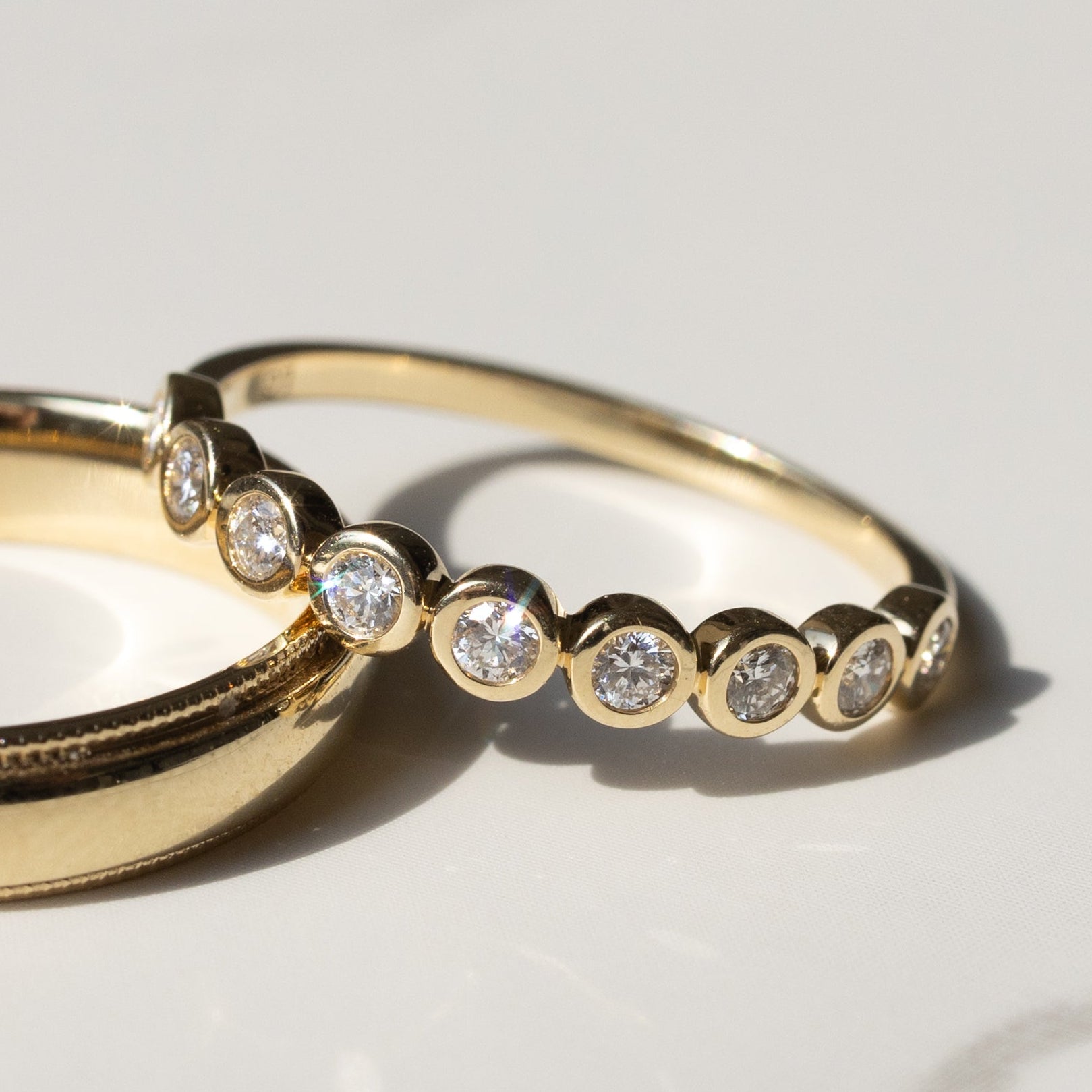 A close up of a gold round diamond bezel wedding band on a soft white marble backdrop with shadows