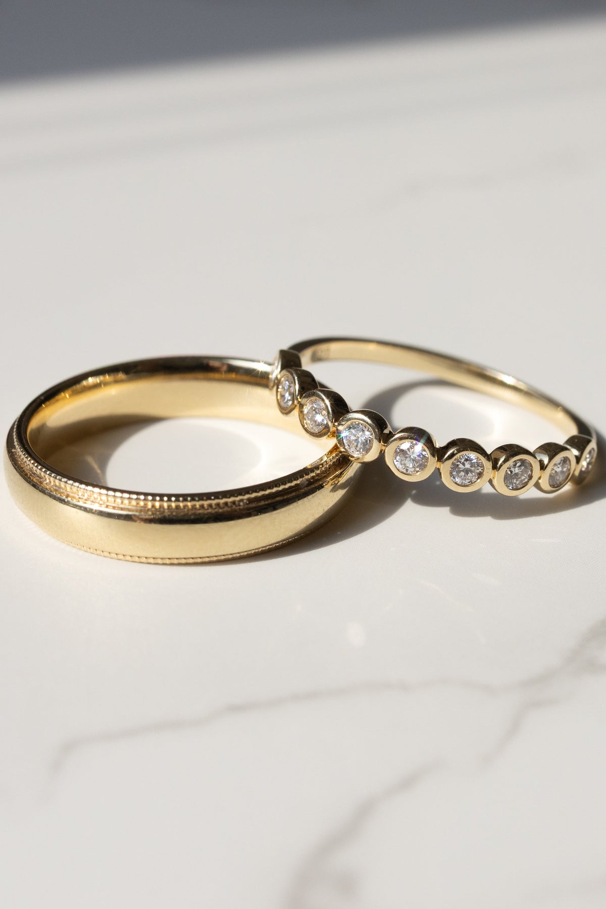 Two gold rings on a white marble surface, one plain and one with a row of small round diamonds.