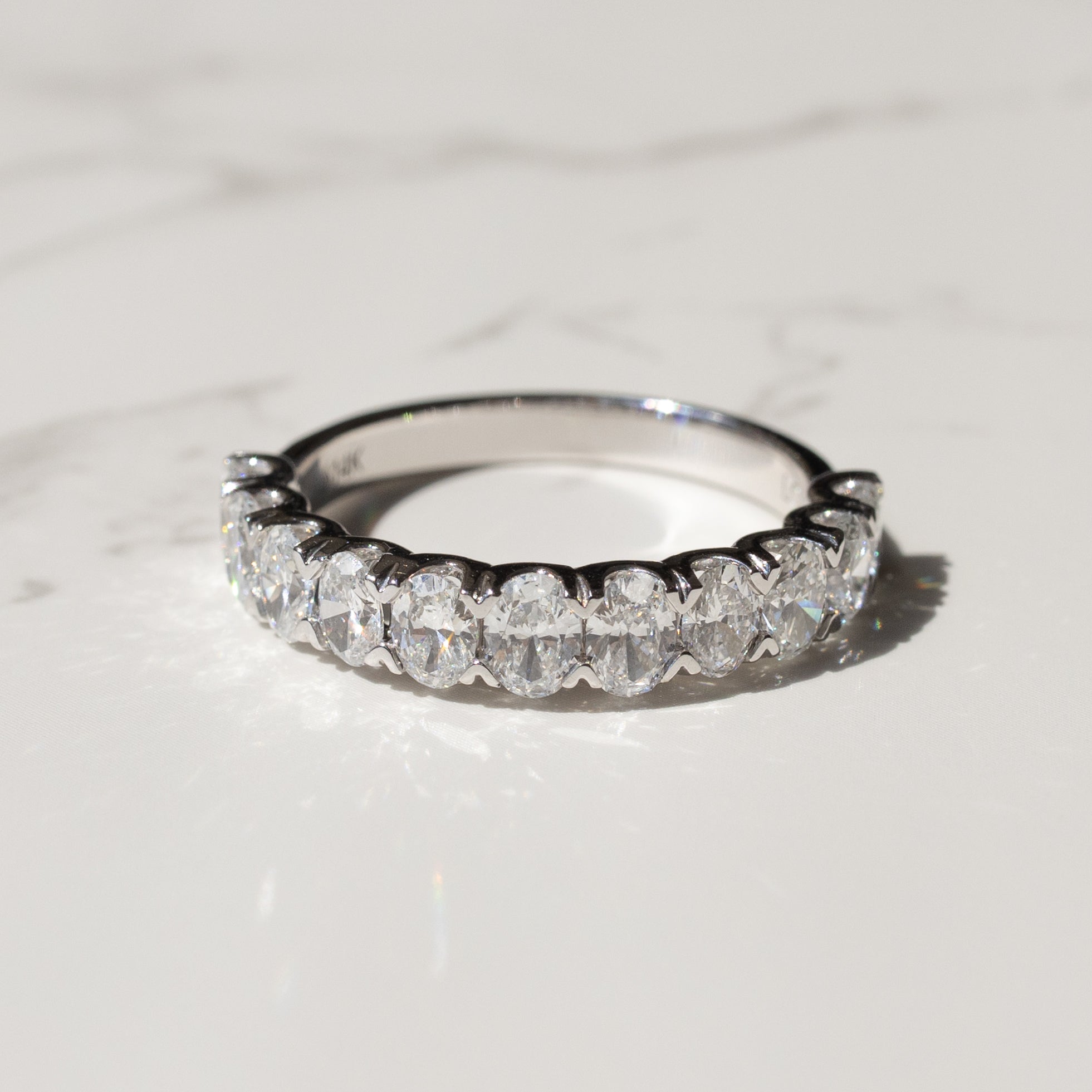A platinum diamond band with large oval diamonds set halfway down the band on a white marble backdrop with shadows