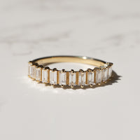 A yellow gold ring featuring a row of baguette-cut diamonds, photographed on a light marble surface with soft shadows.
