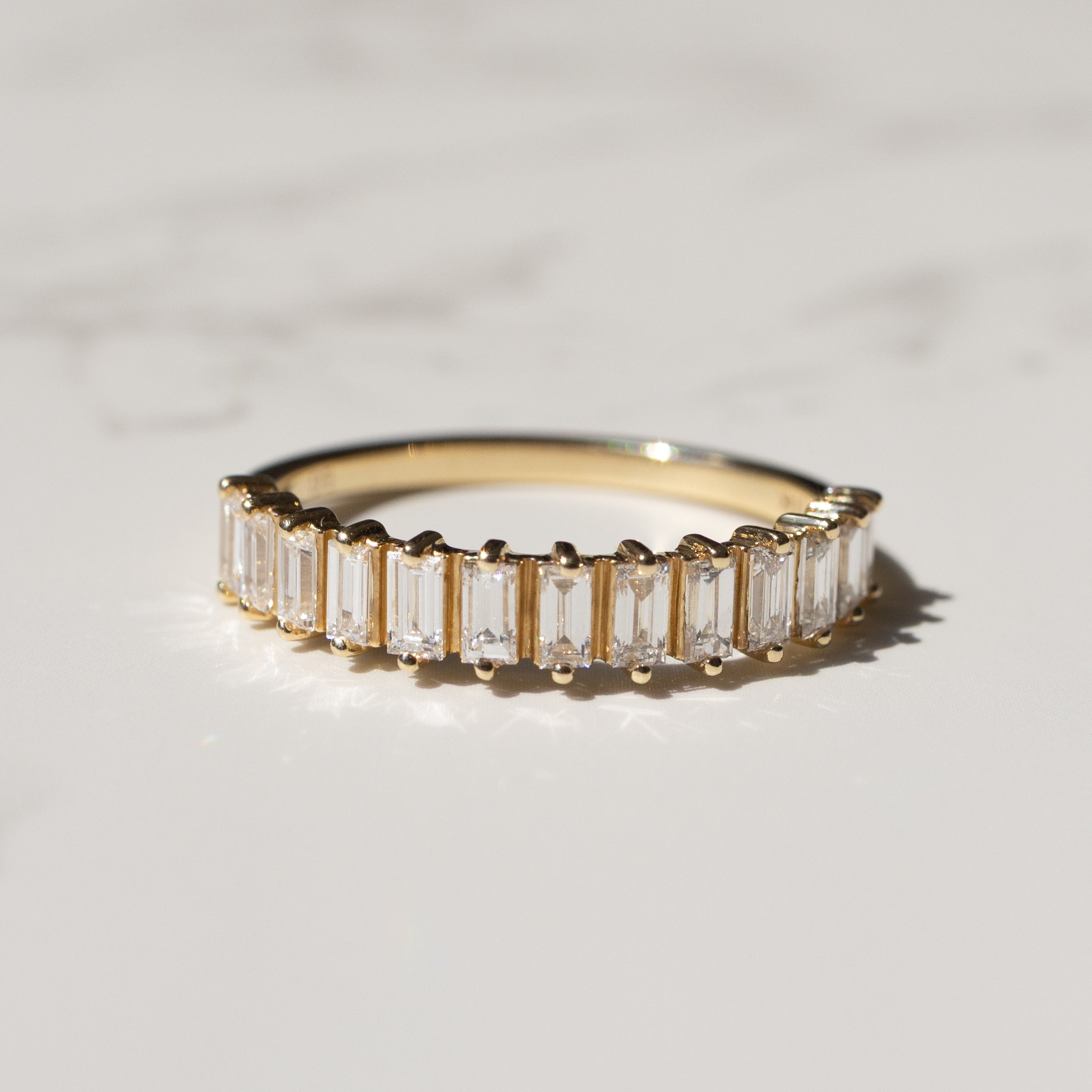 A yellow gold ring featuring a row of baguette-cut diamonds, photographed on a light marble surface with soft shadows.