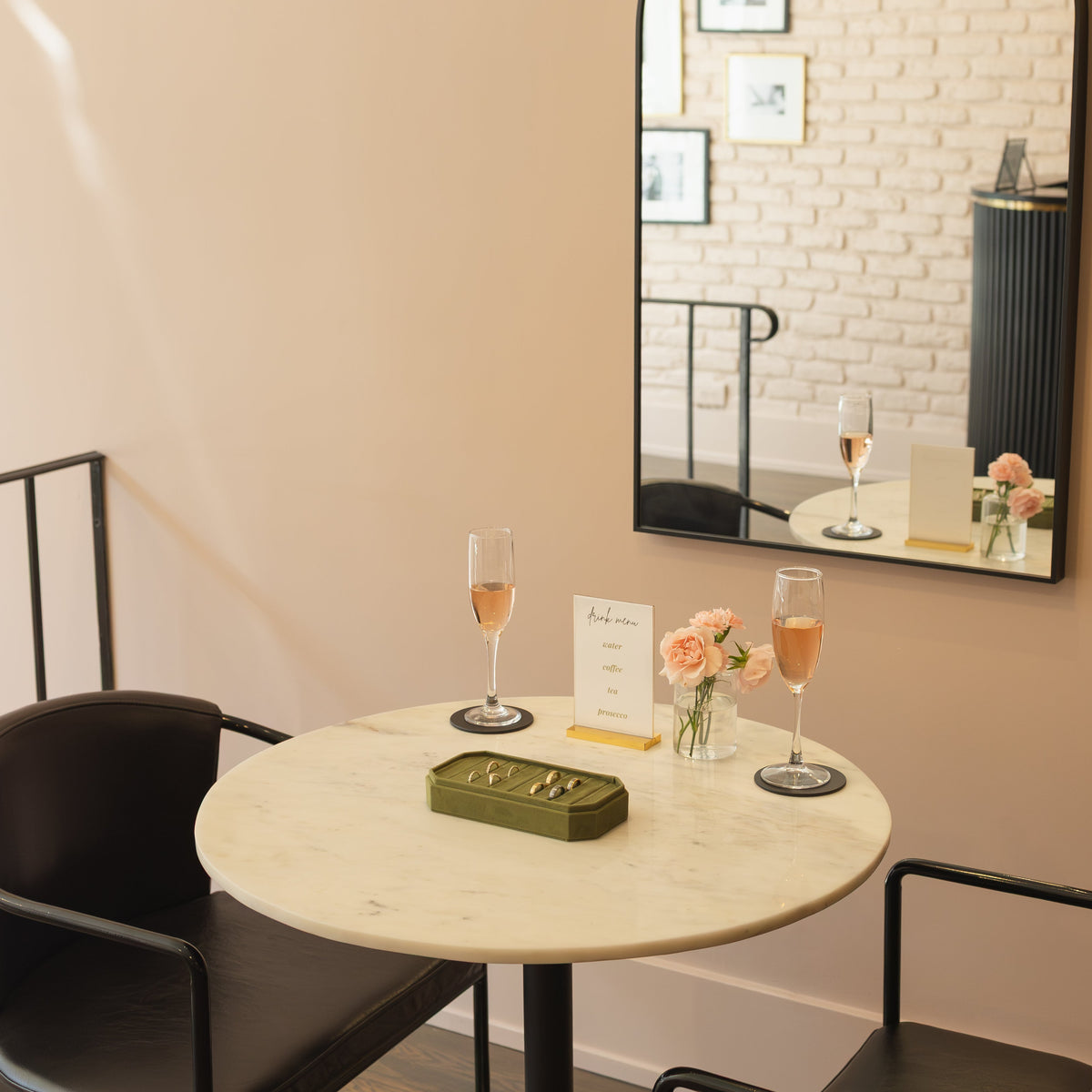 A bistro table with two chairs in a bright showroom. On the bistro table is a green tray with wedding bands, two glasses of champagne, flowers, and a drink menu
