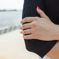 A close-up of a person's hand wearing a thin gold band ring with dainty channel set baguette diamonds, resting on their arm. The person is wearing a black cable-knit sweater, and the background shows a blurred outdoor waterfront scene.