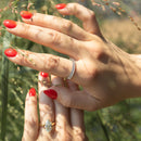 A close-up lifestyle shot of two hands with red nail polish, one wearing a silver oval diamond band ring and the other a gold marquise diamond engagement ring, set against a blurred natural background of green grass.