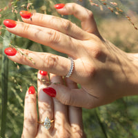 A close-up lifestyle shot of two hands with red nail polish, one wearing a silver oval diamond band ring and the other a gold marquise diamond engagement ring, set against a blurred natural background of green grass.
