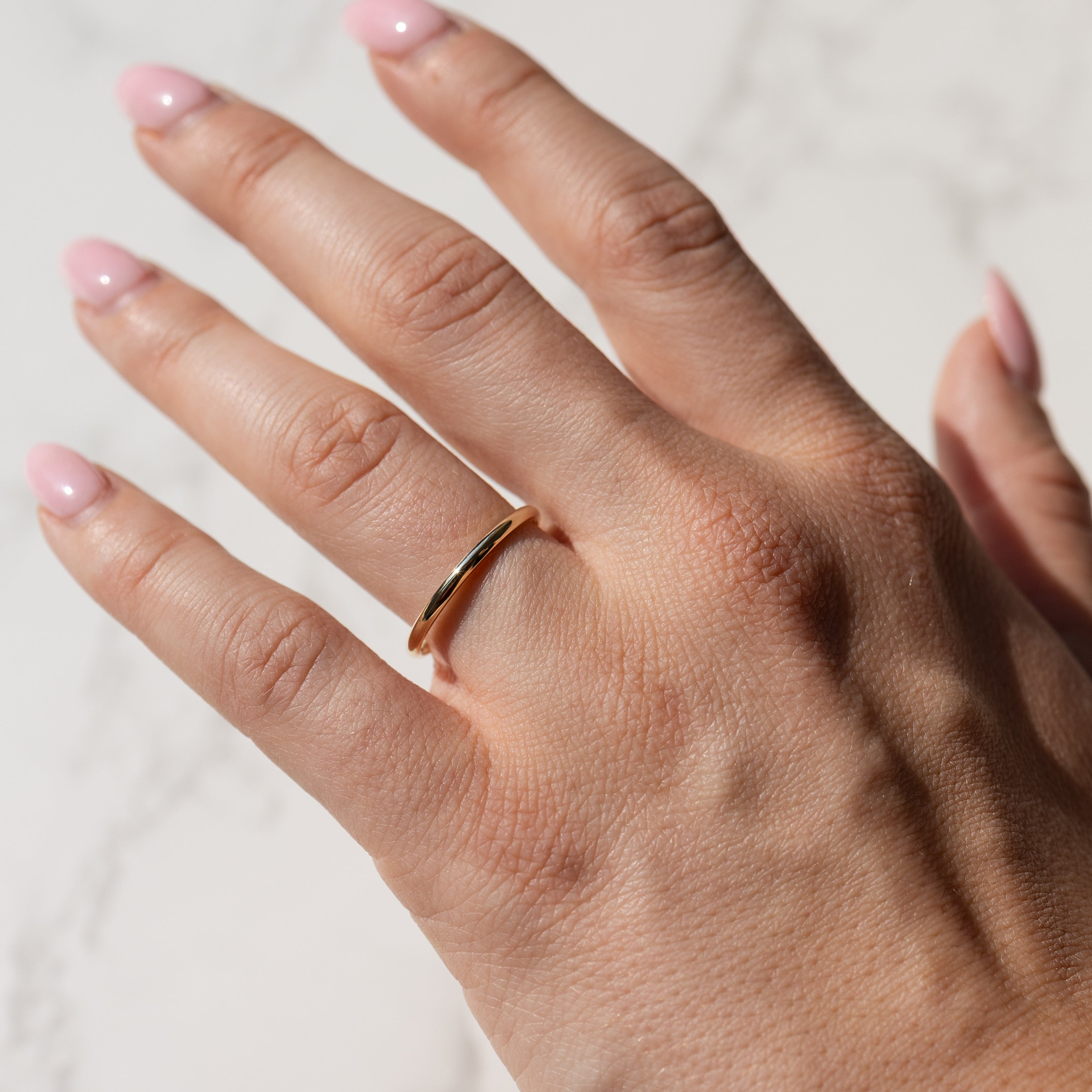 A close-up of a hand wearing a thin, polished gold band ring, set against a light marble background.
