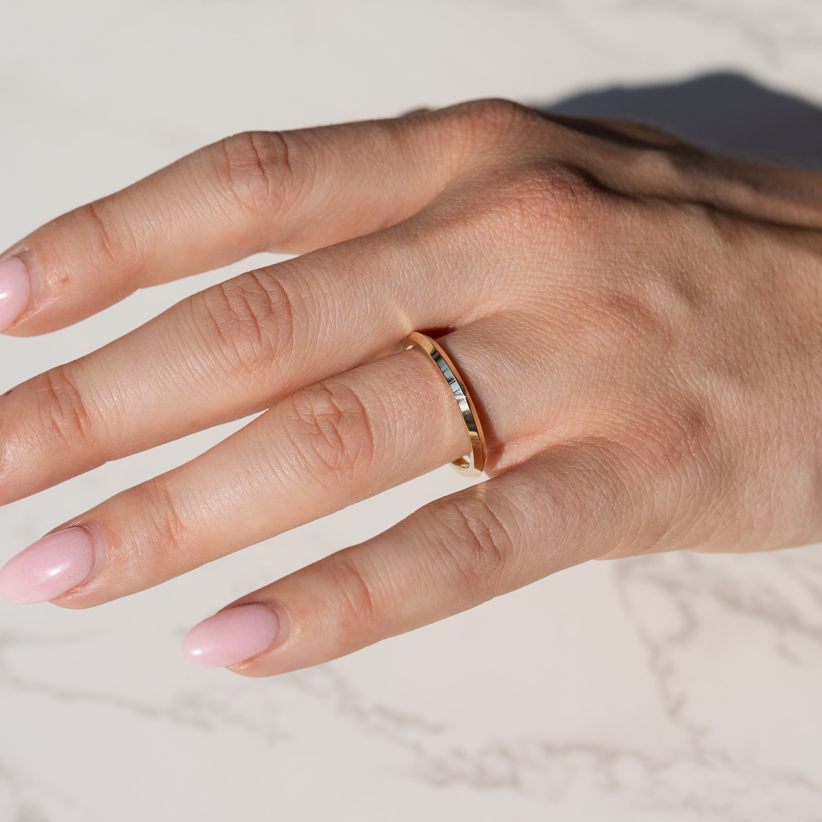 A close-up of a hand wearing a slim gold band ring with a knife edge point along the outer edge, resting on a light marble surface.