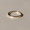 A minimalist gold flat band ring with a polished finish, resting on a neutral beige surface with a soft shadow.