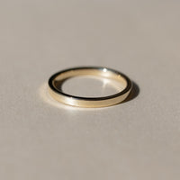 A minimalist gold flat band ring with a polished finish, resting on a neutral beige surface with a soft shadow.