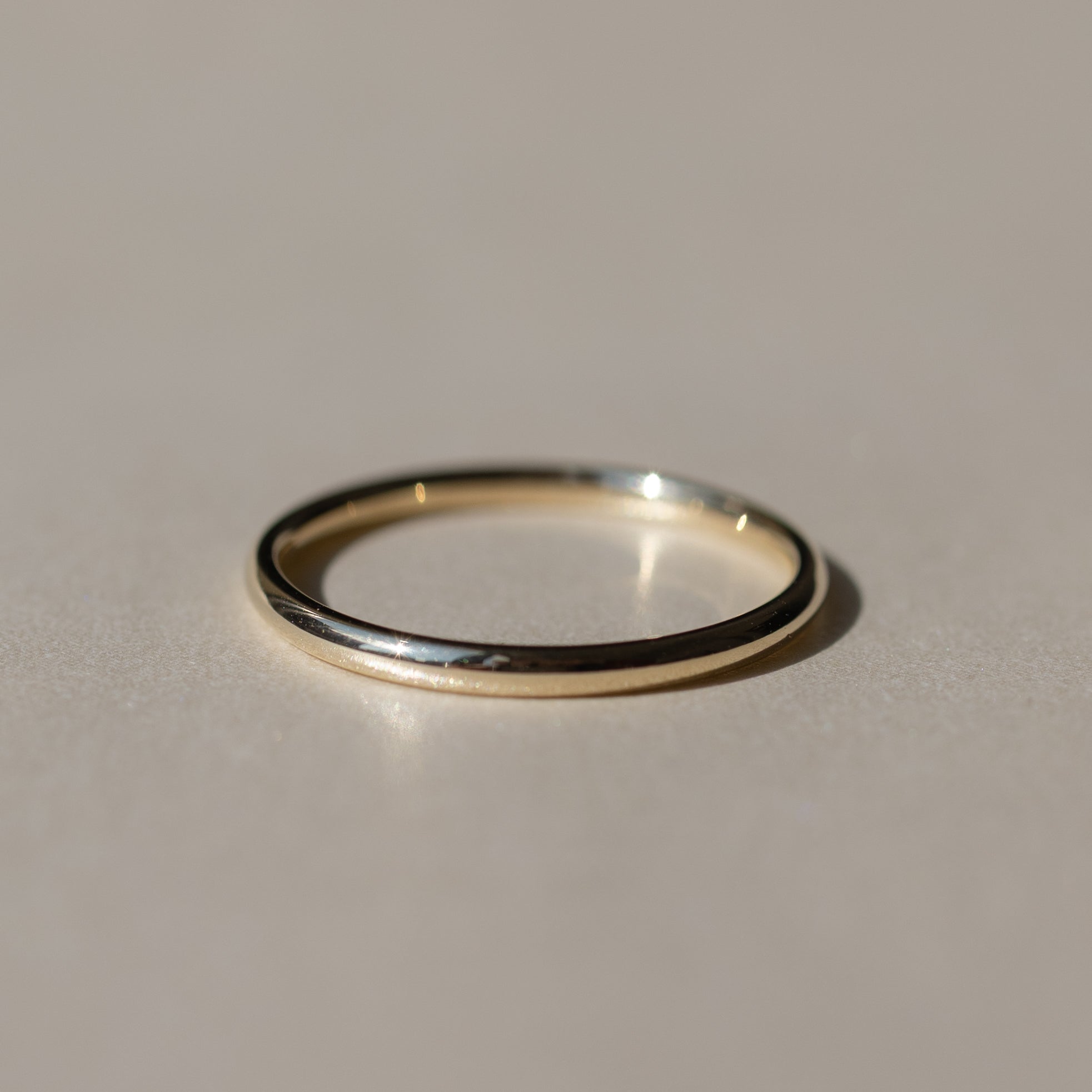 A minimalist, thin gold band ring with a polished texture, resting on a neutral beige surface with a soft shadow.