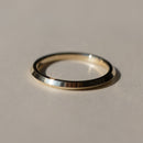 A minimalist polished metal ring, with a knife edge point in the middle of the band, resting on a neutral beige surface with a soft shadow.