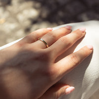 A close-up of a hand wearing a thin gold band ring with a flat face, resting on a textured cream fabric under natural sunlight.