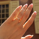 A gold band ring worn on a person's ring finger, shown in warm sunlight against a blurred brick building background.