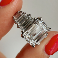 Emerald and baguette platinum diamond ring held by a hand with red nail polish on a neutral background