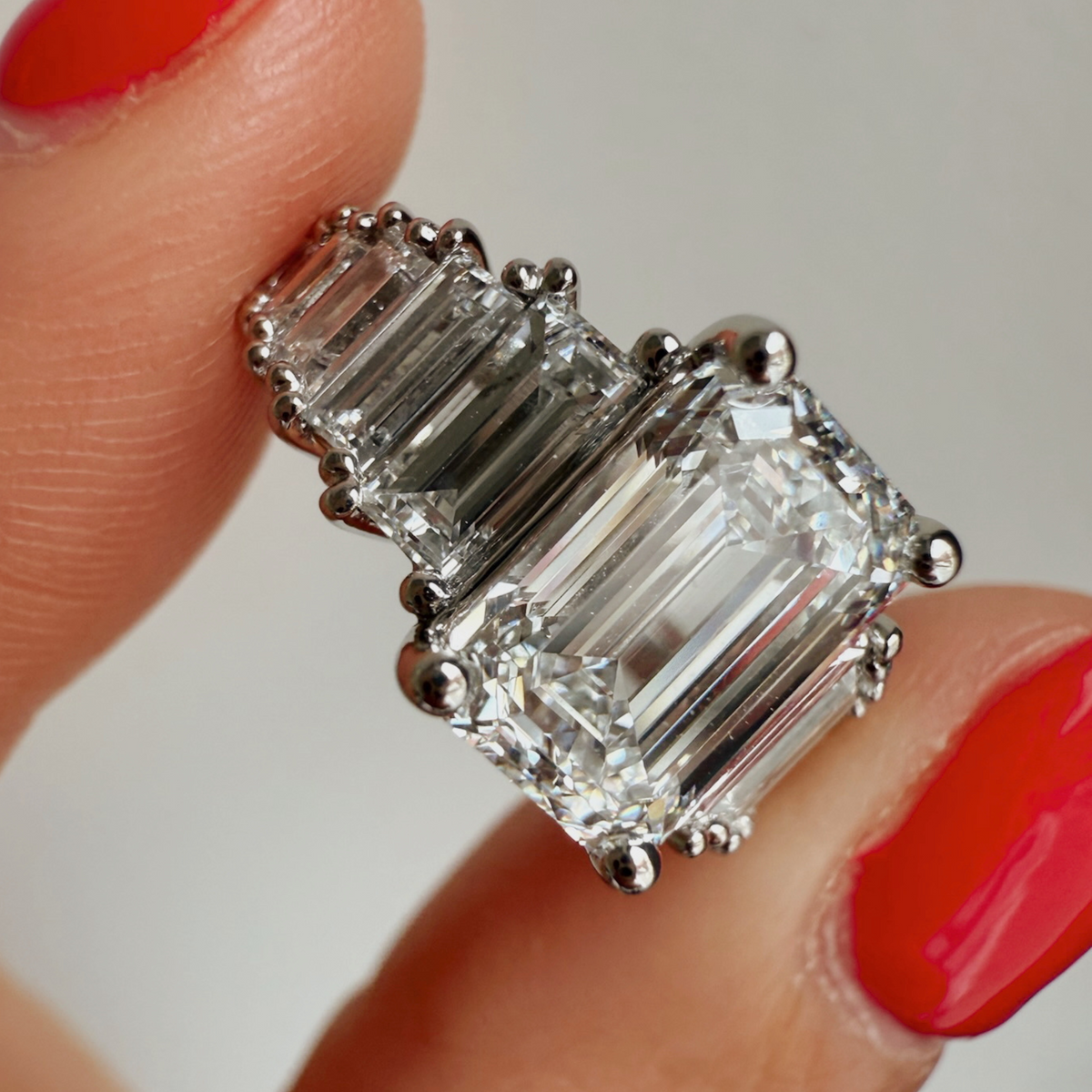 Emerald and baguette platinum diamond ring held by a hand with red nail polish on a neutral background