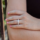 A close-up of a person's hand resting on their arm, wearing a white gold ring set with oval diamonds. The background shows a soft, blurred outdoor scene with green foliage and a dark railing.