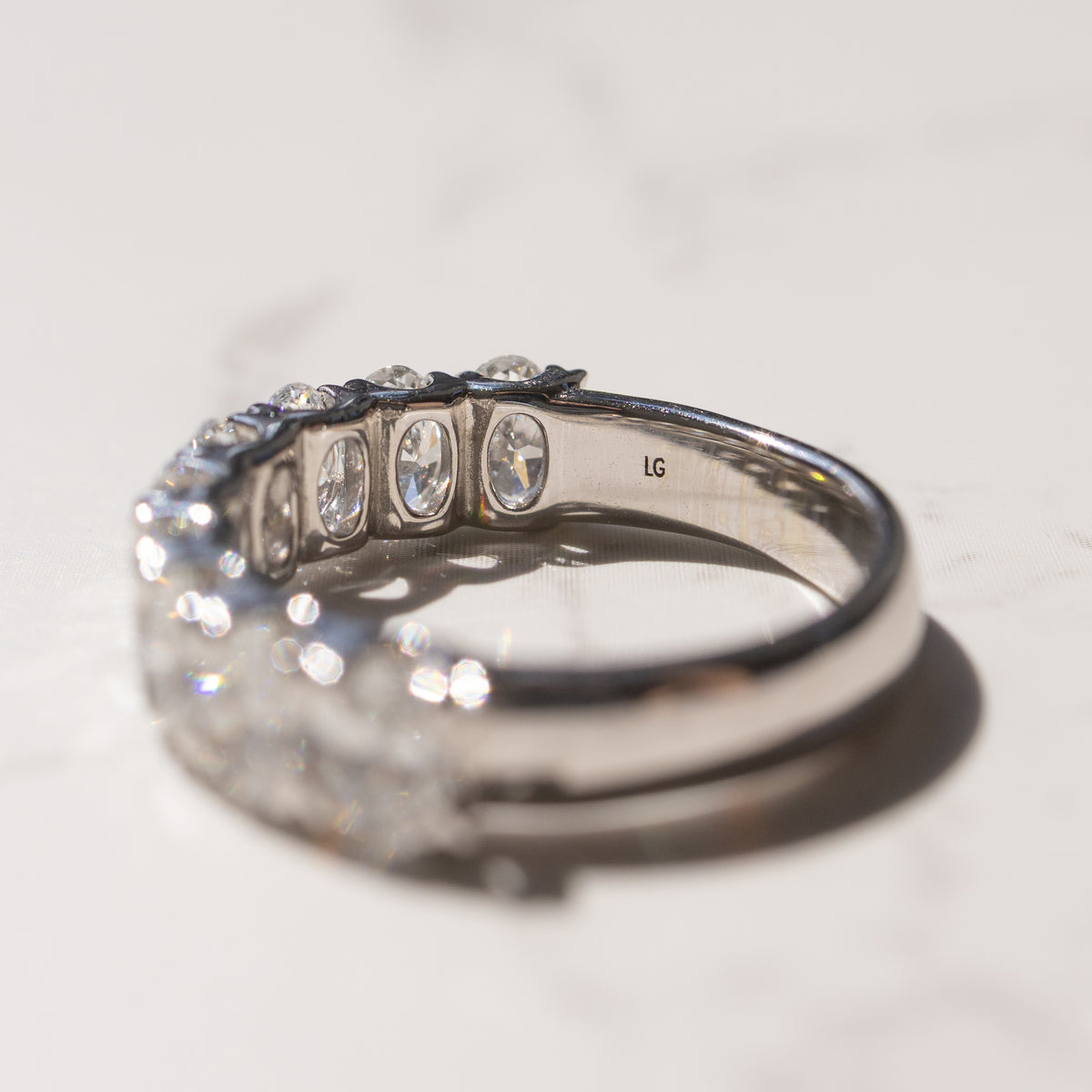 A diamond ring with oval-cut diamonds, shown in a close-up side view on a light marble surface. The inside of the band is stamped with 'LG'.