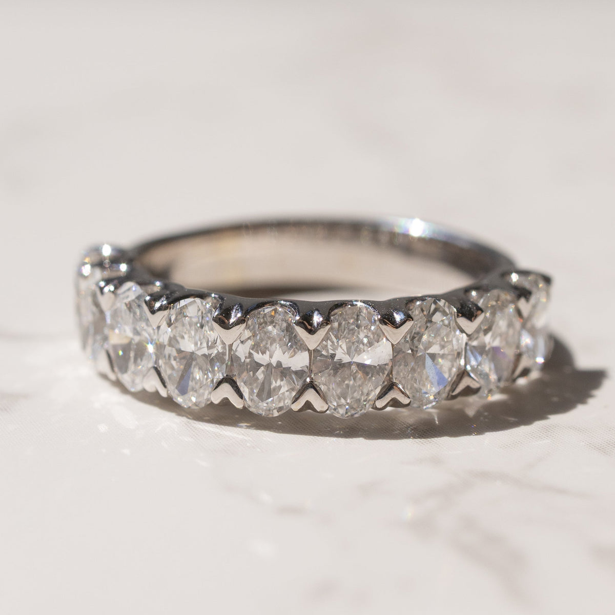 A white gold diamond eternity ring with oval-cut diamonds resting on a light marble surface.