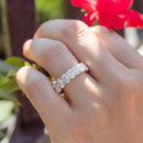 A hand holding a fuchsia colored flower. On the left ring finger of the hand is a very large oval-cut diamond eternity wedding band.