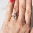 A close-up of a hand wearing a gold ring with a marquise-cut diamond. The ring is shown on the ring finger against a light, neutral background.