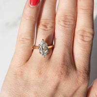 A close-up of a hand wearing a gold ring with a marquise-cut diamond. The ring is shown on the ring finger against a light, neutral background.