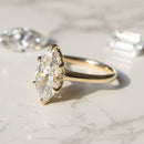 A yellow gold ring with a marquise-cut diamond center stone, displayed on a light marble surface with two loose diamonds in the background.