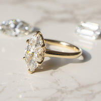 A yellow gold ring with a marquise-cut diamond center stone, displayed on a light marble surface with two loose diamonds in the background.