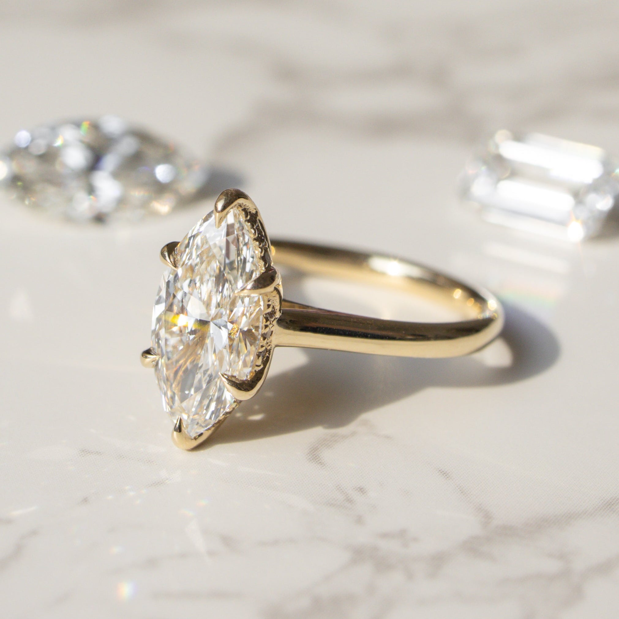 A yellow gold ring with a marquise-cut diamond center stone, displayed on a light marble surface with two loose diamonds in the background.