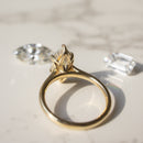 A gold ring with a marquise-cut gemstone in a tulip prong setting with a hidden halo, displayed on a light marble surface with two loose diamonds nearby.