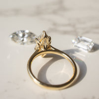 A gold ring with a marquise-cut gemstone in a tulip prong setting with a hidden halo, displayed on a light marble surface with two loose diamonds nearby.