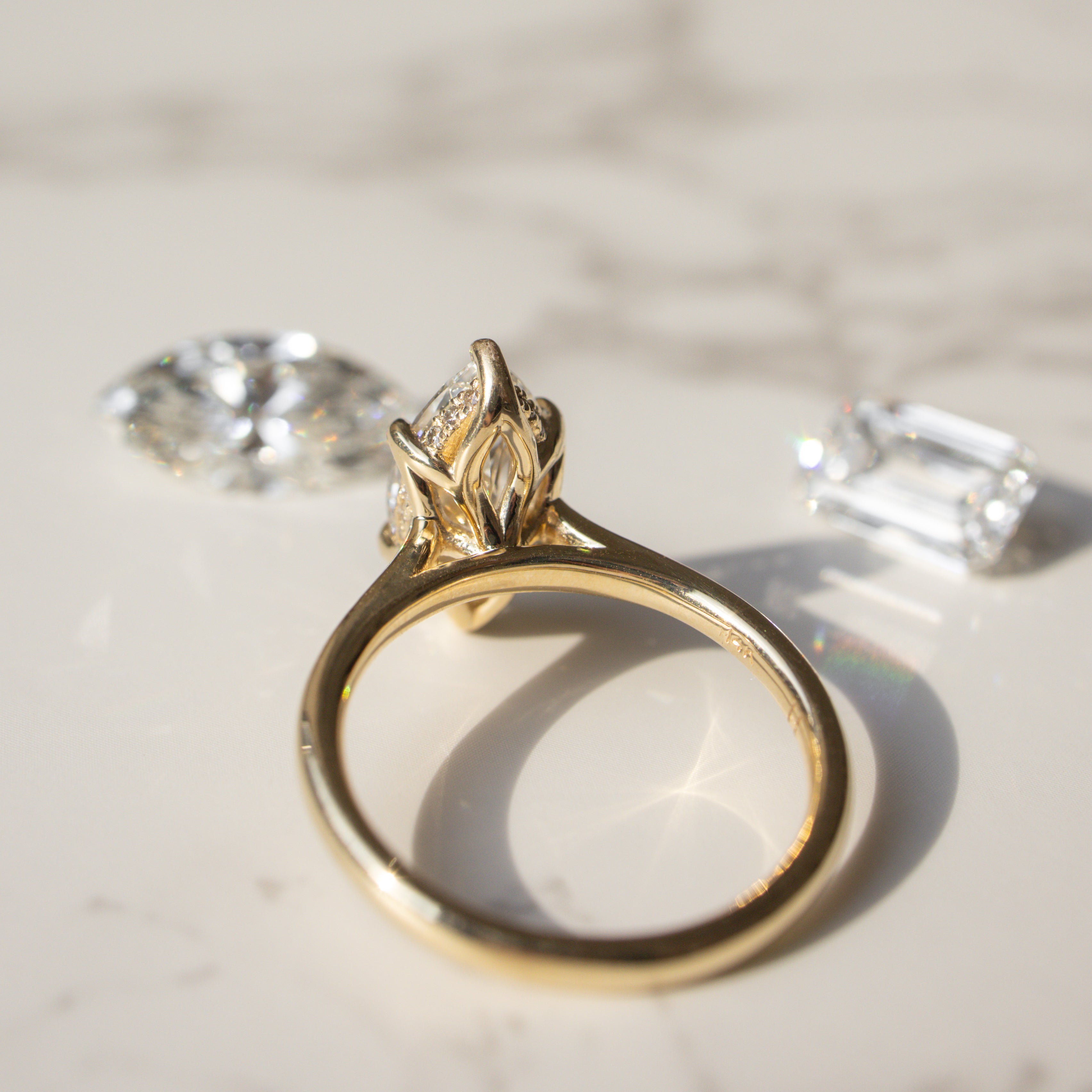 A gold ring with a marquise-cut gemstone in a tulip prong setting with a hidden halo, displayed on a light marble surface with two loose diamonds nearby.