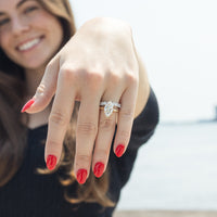A close-up of a woman's hand wearing a dual-toned bridal ring stack with a marquise-cut center diamond and smaller oval stones, photographed outdoors with a blurred bright sky and water background.