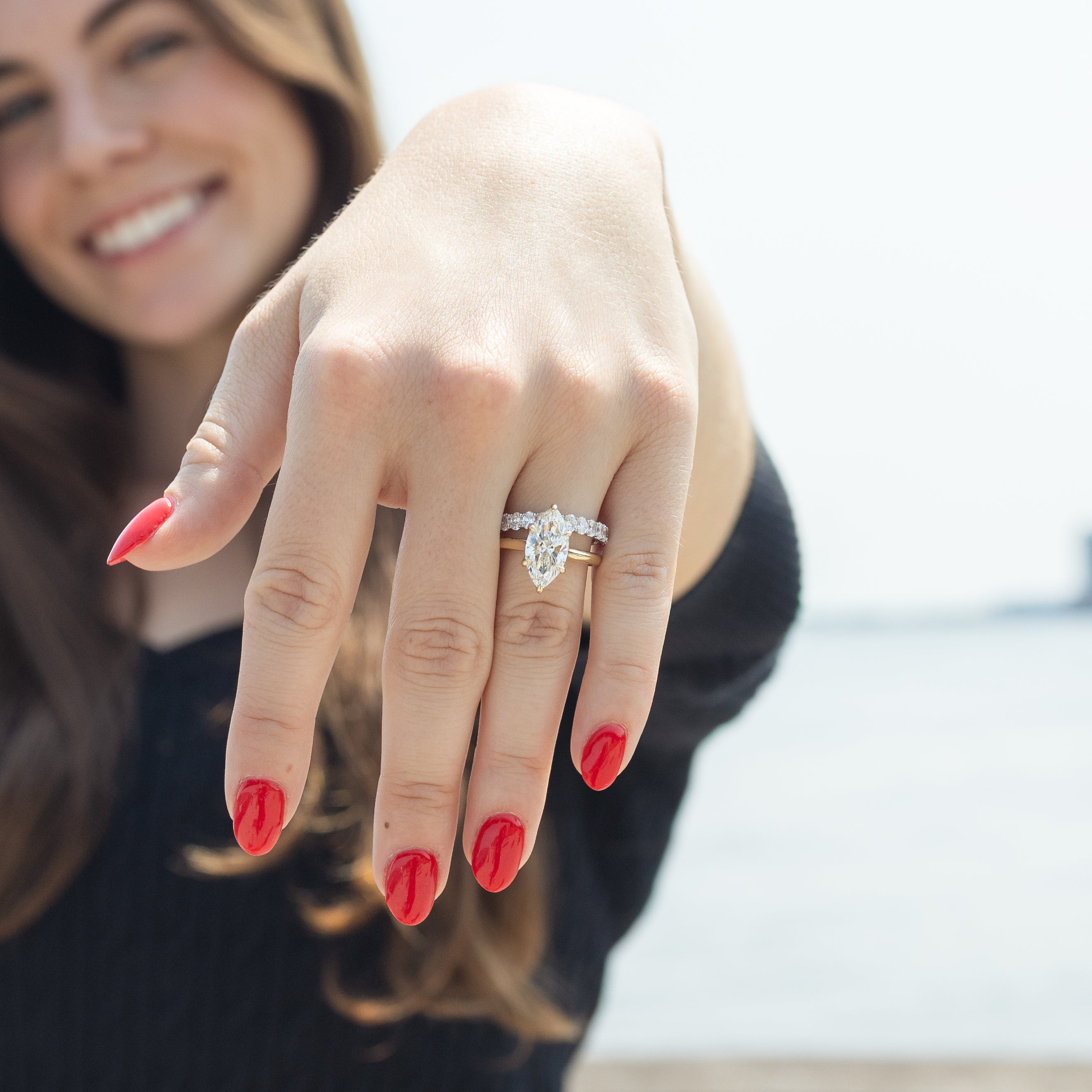 A close-up of a woman's hand wearing a dual-toned bridal ring stack with a marquise-cut center diamond and smaller oval stones, photographed outdoors with a blurred bright sky and water background.