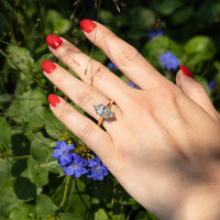 A close-up of a hand with bright red nail polish wearing a gold ring with a marquise-cut clear diamond, set against a background of green foliage and small blue flowers.
