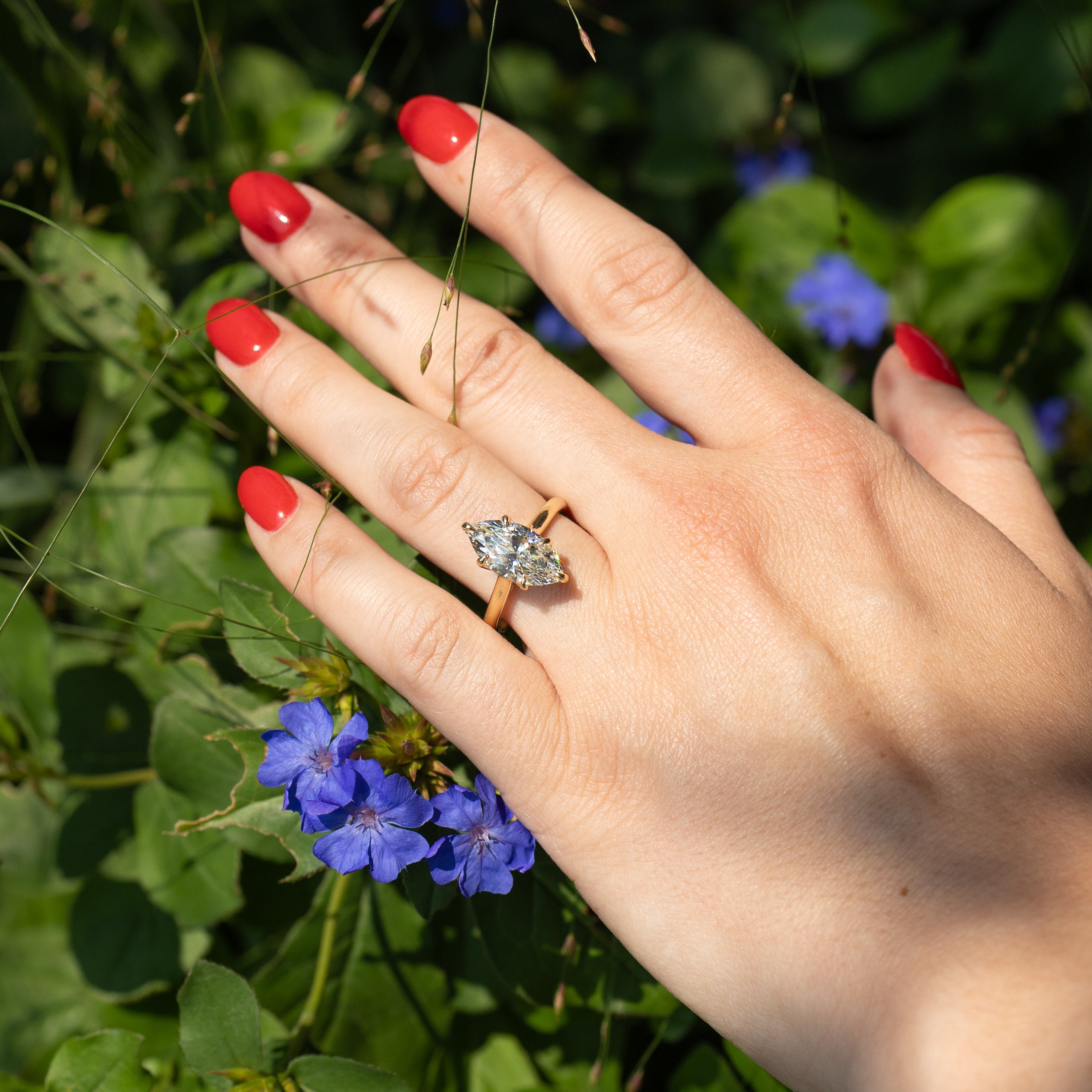 A close-up of a hand with bright red nail polish wearing a gold ring with a marquise-cut clear diamond, set against a background of green foliage and small blue flowers.