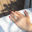 A close-up of a hand with red nail polish resting on white fabric, wearing a gold ring with a large marquise diamond and a wedding band with oval diamonds. The background shows a wooden deck with dappled sunlight.