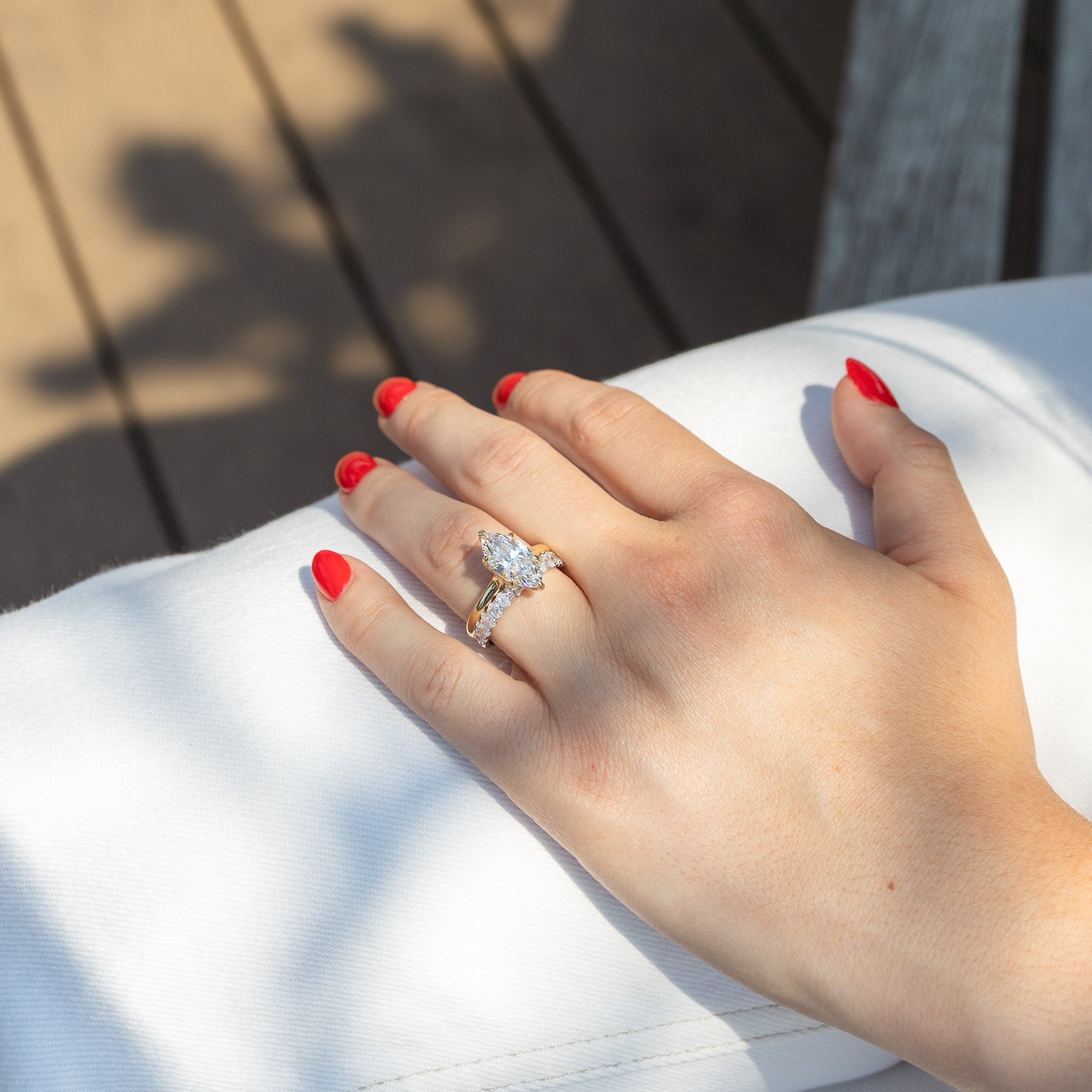 A close-up of a hand with red nail polish resting on white fabric, wearing a gold ring with a large marquise diamond and a wedding band with oval diamonds. The background shows a wooden deck with dappled sunlight.