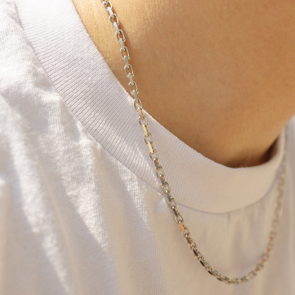 A close-up of a person's neck and upper chest wearing a silver-toned chain necklace over a white t-shirt.