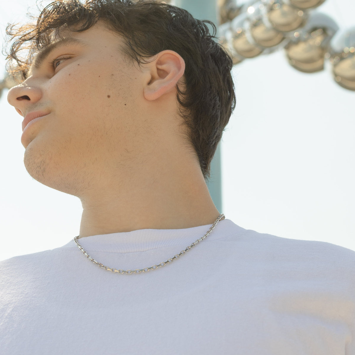A man in a white t-shirt wearing a silver chain necklace, shown in profile against a bright outdoor background with a reflective metallic sphere sculpture.