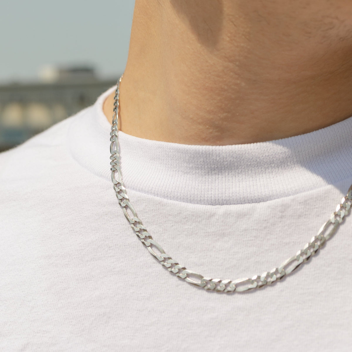 A close-up of a person's neck and upper chest wearing a silver chain necklace with a white t-shirt, set against a blurred outdoor background.