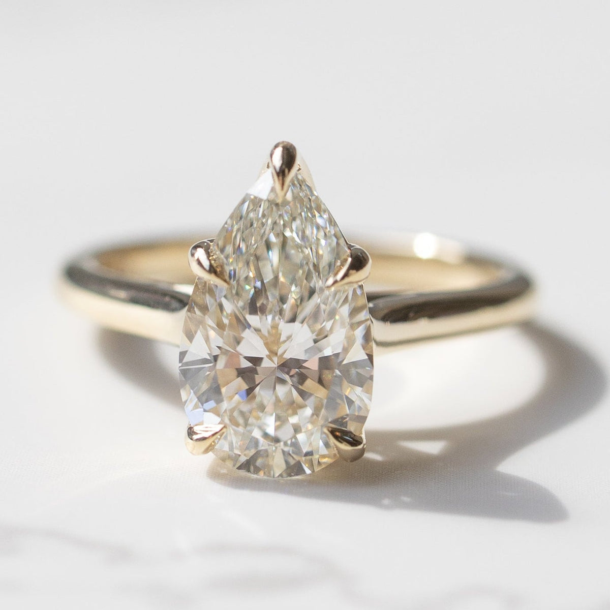 A pear cut engagement ring in a yellow gold setting sitting on a white marble backdrop with soft grey shadows.