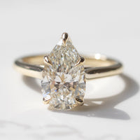 A pear cut engagement ring in a yellow gold setting sitting on a white marble backdrop with soft grey shadows.